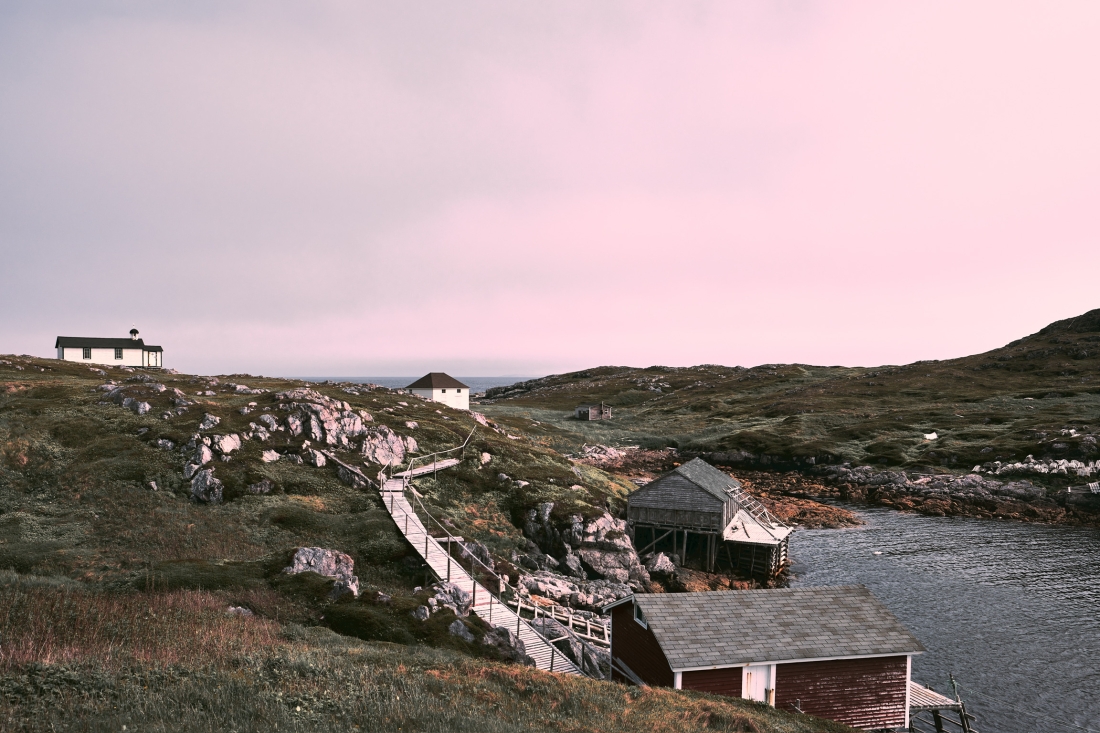 A boardwalk winds down a rocky slope to red fishing stores on a dark inlet, under a pink evening sky.