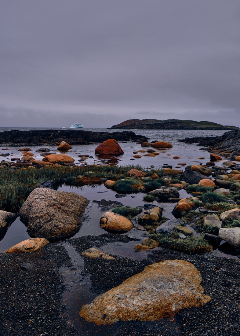 Orange boulders in a tidal pool on a dark shore, with an iceberg and a low island on the horizon.