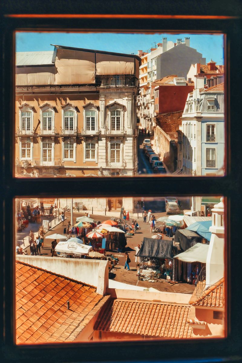 Two window openings cut into a dark stone wall. The upper one frames old Lisbon facades and a narrow street. The lower one frames a street market under umbrellas, above terracotta roofs.