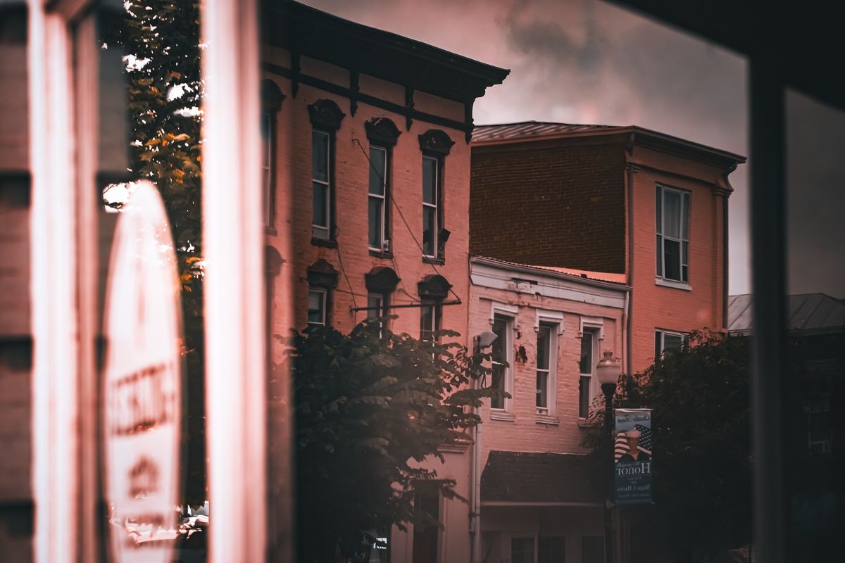 Brick storefronts on a small-town street under a heavy sky, seen through a window that carries the reflection of a sign.