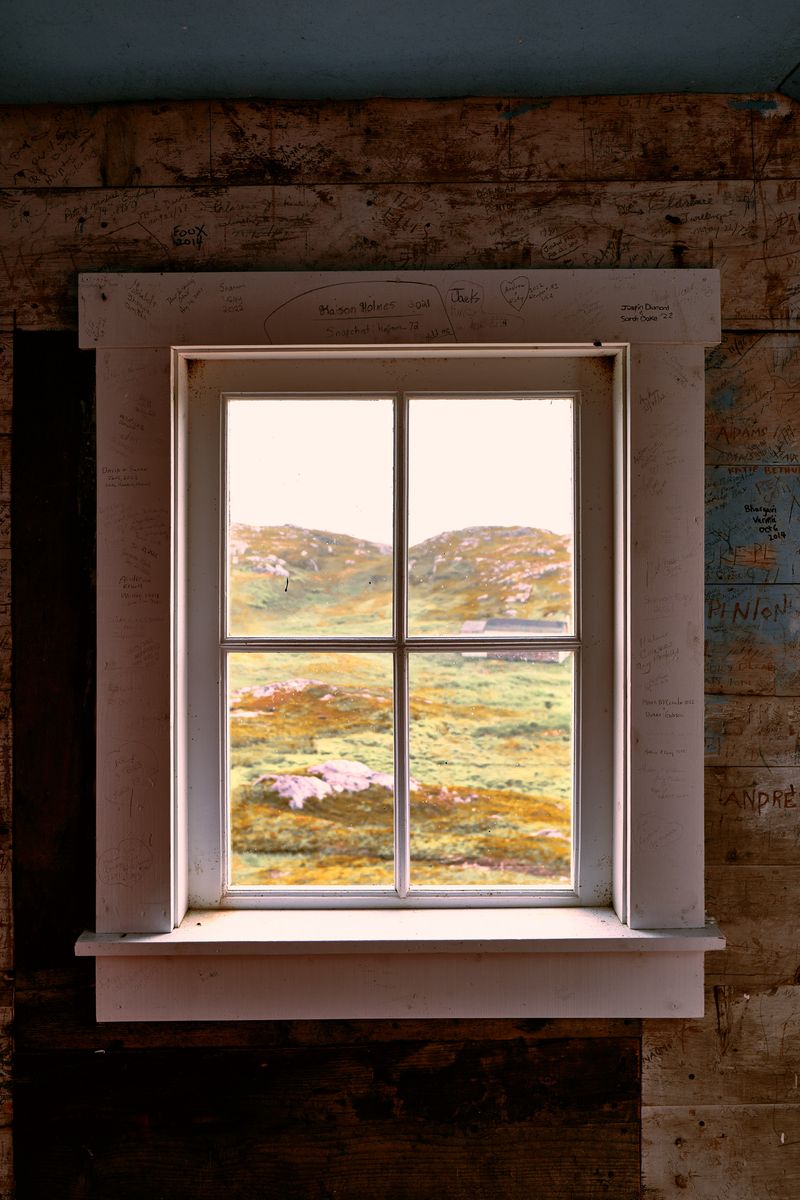 A four-paned window in a wooden church wall covered in visitors' signatures, looking out on green and gold moorland and pale rock.